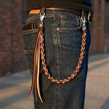 Side view of a model wearing a braided black and brown leather wallet chain with long hanging tassels attached to blue jeans belt loops, featuring silver-tone clasps and a wallet in the back pocket.