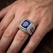 Primo piano di una mano maschile che indossa un grande anello a croce gotica con un accento di pietra blu, catturato in una morbida illuminazione da studio.