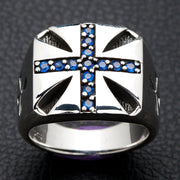 A solid sterling silver Maltese cross ring, inlaid with brilliant blue stones, shown on a dark, textured background.
