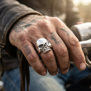 粗いタトゥーのある手が、ダイヤモンドの石が一つの目のくぼみにセットされた磨かれたスターリングシルバーのスカルリングを着けて、バイクのハンドルをしっかりと握っているクローズアップ。