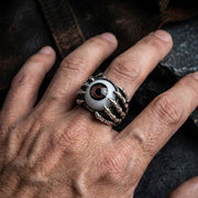 Close-up de uma mão a usar um anel gótico de garra com olho prateado, com um centro de olho realista segurado por garras, fotografado em iluminação sombria.