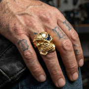 Žlutý zlatý prsten ve tvaru lebky s modrým CZ okem, nošený na tetované ruce, ukazující podrobnou řemeslnou práci a značnou velikost.