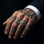 Sterling silver sugar skull ring featuring green gemstone eyes worn on a tattooed male hand, showcasing detailed biker jewelry design.