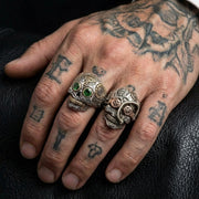 Sterling silver sugar skull ring featuring green gemstone eyes worn on a tattooed male hand, showcasing detailed biker jewelry design.