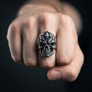 Närbild av en hand som bär en ring i sterlingsilver med en inre demon skalle, med detaljerad gotisk skalle ansiktsdesign mot en mörk suddig bakgrund.