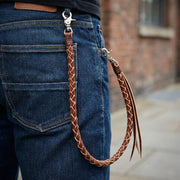 Corrente de carteira de couro trançado castanho claro com fechos de lagosta prateados ligados ao bolso traseiro de jeans azul escuro, apresentando franjas decorativas de couro num ambiente de estilo urbano.