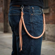 Lichtbruine gevlochten leren portemonneeketting bevestigd aan een donkerblauwe jeans, waarbij de natuurlijke valling van de riemlus naar de achterzak zichtbaar is.