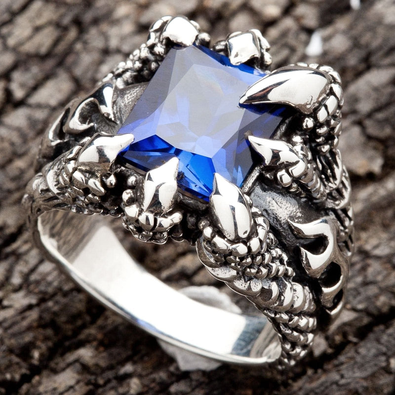 Cette bague pour homme en argent massif présente quatre griffes monstrueuses serrant une pierre massive en saphir bleu CZ de taille carrée.