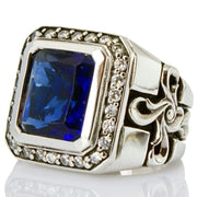 A men's solid sterling silver gothic cross ring, featuring a large blue sapphire CZ stone and a halo of clear accents.