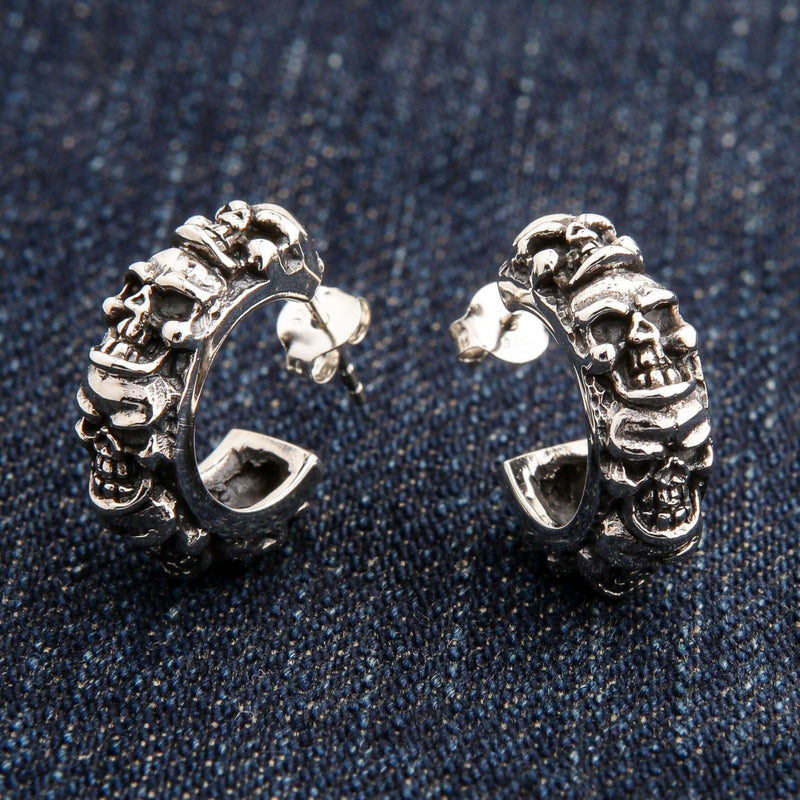 Ein Paar Herren-Ohrringe aus Sterlingsilber mit mehreren Totenköpfen, Steckverschluss und Schmetterlingsverschluss, auf einer blauen Jeans liegend.
