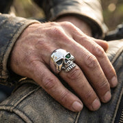 特寫一隻男性手,戴著一枚拋光的純銀骷髏戒指,戒指上有一顆綠色寶石眼睛,手放在一件磨損的棕色皮革摩托車夾克上。