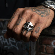 Silver MF Doom mask ring worn on middle finger of tattooed hand making a fist, featuring iconic villain helmet design with eye holes and angular metal craftsmanship, urban graffiti background with luxury watch visible on wrist
