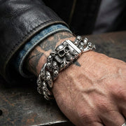 Heavy silver multi-skull Cuban link bracelet with detailed skull clasp worn on tattooed male wrist with black leather jacket sleeve, showcasing edgy biker jewelry style