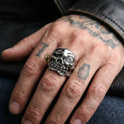 Silver pirate skull ring with crystal gemstone eye and stitched mouth detail worn on tattooed hand resting on denim jeans with black leather jacket, biker jewelry lifestyle shot