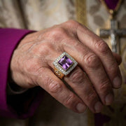 優雅的公主方切割紫水晶主教戒指,鑲嵌於金色的鑽石光環中,佩戴在一位神職人員的手上,背景中可見紫色的祭服袖子和銀色十字架。