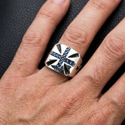 خاتم صليب مالطي فضي مع أحجار زرقاء يُرتدى على الإصبع الأوسط ضد خلفية مظلمة ذات نسيج تُظهر تصميم الصليب المعقد وترتيب بافي.