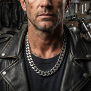 Thick silver Cuban chain necklace worn by tattooed man in black leather jacket standing in motorcycle workshop with tools