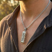 Man wearing a sterling silver mini harmonica pendant with intricate floral engraving on a chunky curb chain necklace, styled with an open dark shirt in warm natural lighting