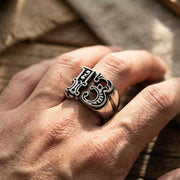 Ornate sterling silver number 13 ring with decorative gothic styling worn on a man's finger, featuring raised numerals and scrollwork design against a rustic wooden background