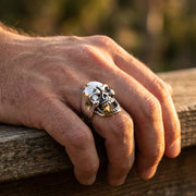 Stříbrný prsten s kouřící lebkou s diamantovým okem a zlatými akcenty, nošený na mužské ruce opřené o zvětralé dřevo, s detailními zuby a designem v motorkářském stylu.
