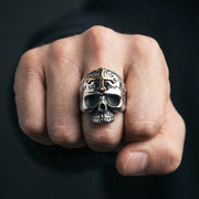 Närbild av en tung ring i sterlingsilver med ett dödskallemotiv som har en guldkorsaccent på pannan och ingraverad text, bärs på en mans knutna näve mot en mörk bakgrund.