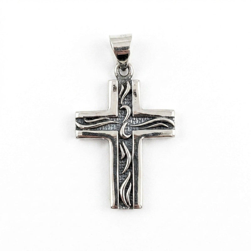 Vy framifrån av ett litet sterlingsilver korshänge med etsade tribal-mönsterlinjer på en texturerad mörk bakgrund
