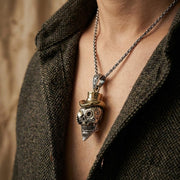 Lähikuva elämäntyyliotoksesta miehestä, joka käyttää yksityiskohtaista sterlinghopeista sokerikallo-riipusta, jossa on kontrastina messinkinen silinterihattu ja kaiverretut kasvosuunnitelmat, tyyliteltynä teksturoidulla tummalla tweed-takilla.