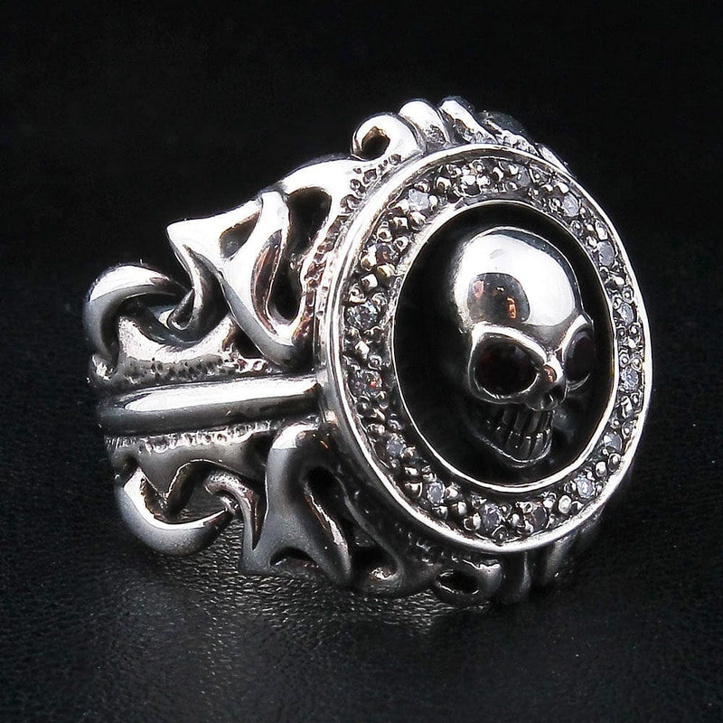 Anillo de plata con calavera tribal y ojos de granate en ángulo de tres cuartos; la calavera rodeada por un halo de circonita transparente sobre una banda calada con volutas.