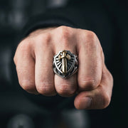 Lähikuva kädestä, jossa on kaksivärinen hopea- ja kultainen ritarisuoja-sormus, jossa on kohotettu kultainen miekka-embleemi, valokuvattu tunnelmallisessa valaistuksessa tumman sumealla taustalla.