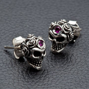 A pair of sterling silver rose skull earrings with brilliant red stone accents shown on a black leather surface.
