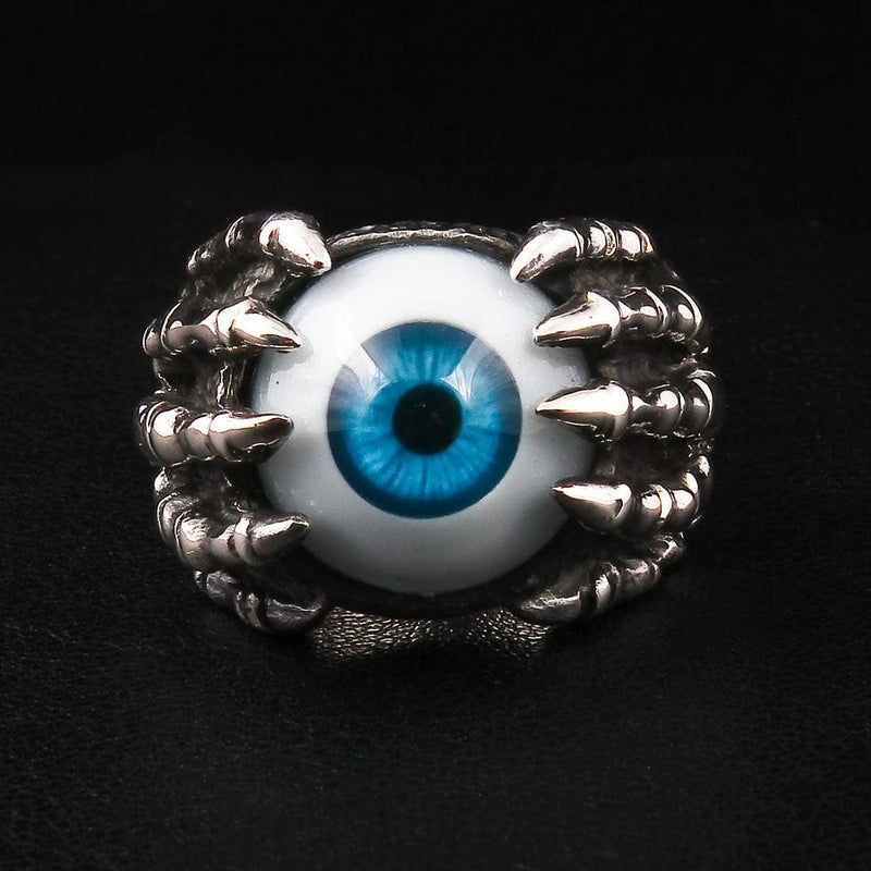 Un anillo de garra de ojo de plata esterlina con un ojo azul realista sostenido por una mano de esqueleto, mostrado sobre un fondo negro.
