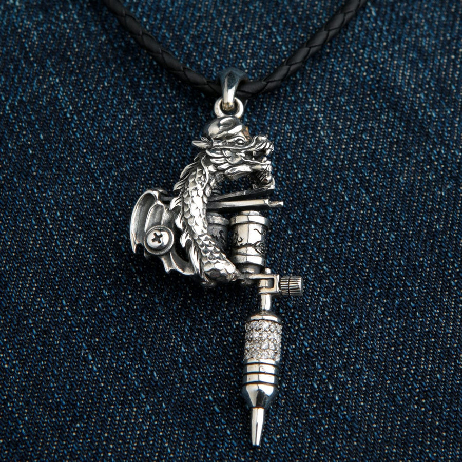 Pendentif en argent sterling représentant une machine à tatouer un dragon avec une pierre scintillante, monté sur un cordon en cuir tressé noir sur fond de denim.