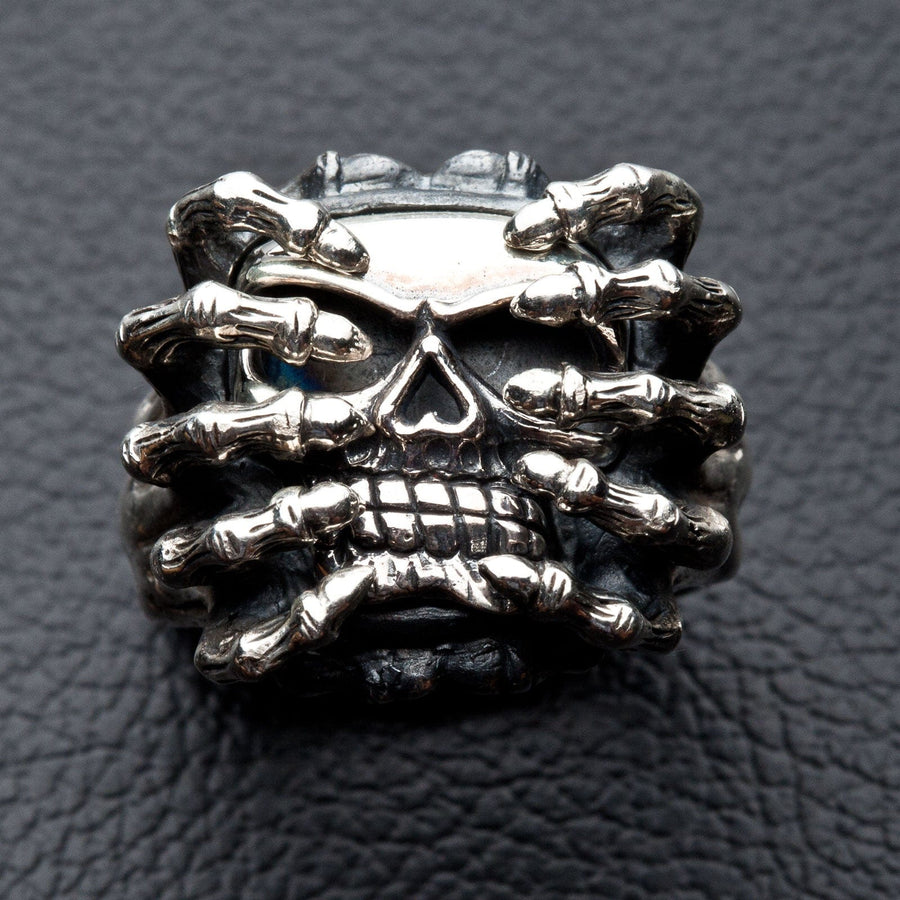 Vista frontal del anillo de calavera de araña de plata esterlina pesada, mostrando la calavera agarrada por manos esqueletales.