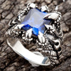 Um anel masculino em prata esterlina sólida apresentando quatro garras monstruosas agarrando uma enorme pedra de zircônia cúbica azul em corte quadrado.