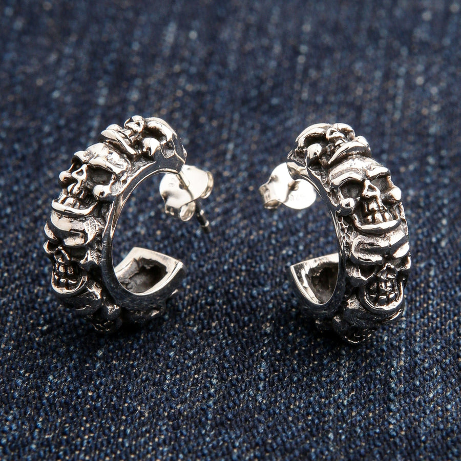 Une paire de boucles d'oreilles pour homme en argent sterling avec plusieurs crânes, avec un poteau et un dos papillon, reposant sur un blue-jean.