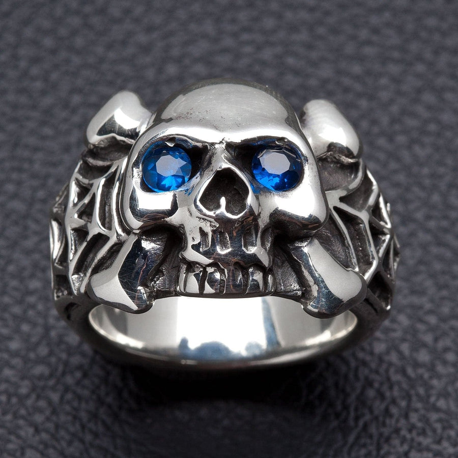 Anillo de calavera de araña de plata 925 maciza con ojos azules y huesos cruzados sobre fondo negro.