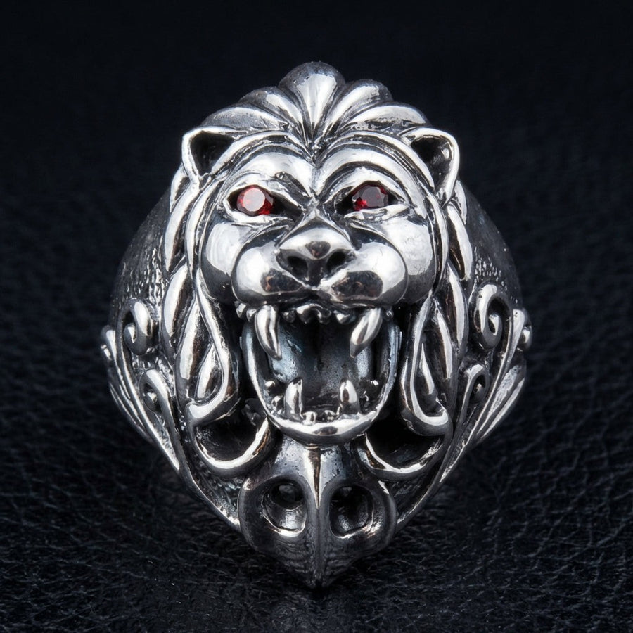 Anillo de cabeza de león en plata de ley con ojos de granate rojo, vista frontal que muestra el rostro del león rugiendo y una melena detallada.