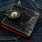 A men's hand-carved black tooled leather wallet with a 925 sterling silver and turquoise snap, shown closed on blue jeans.