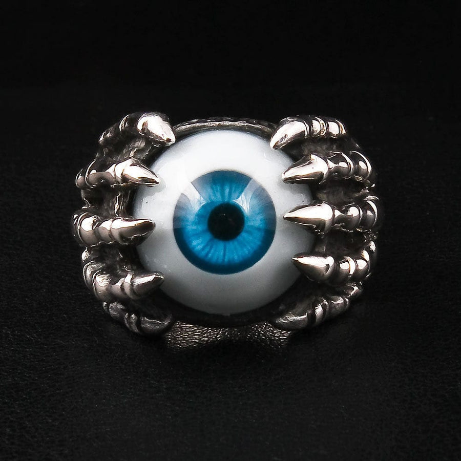 Um anel de garra de olho em prata esterlina com um olho azul realista segurado por uma mão de esqueleto, apresentado sobre um fundo preto.